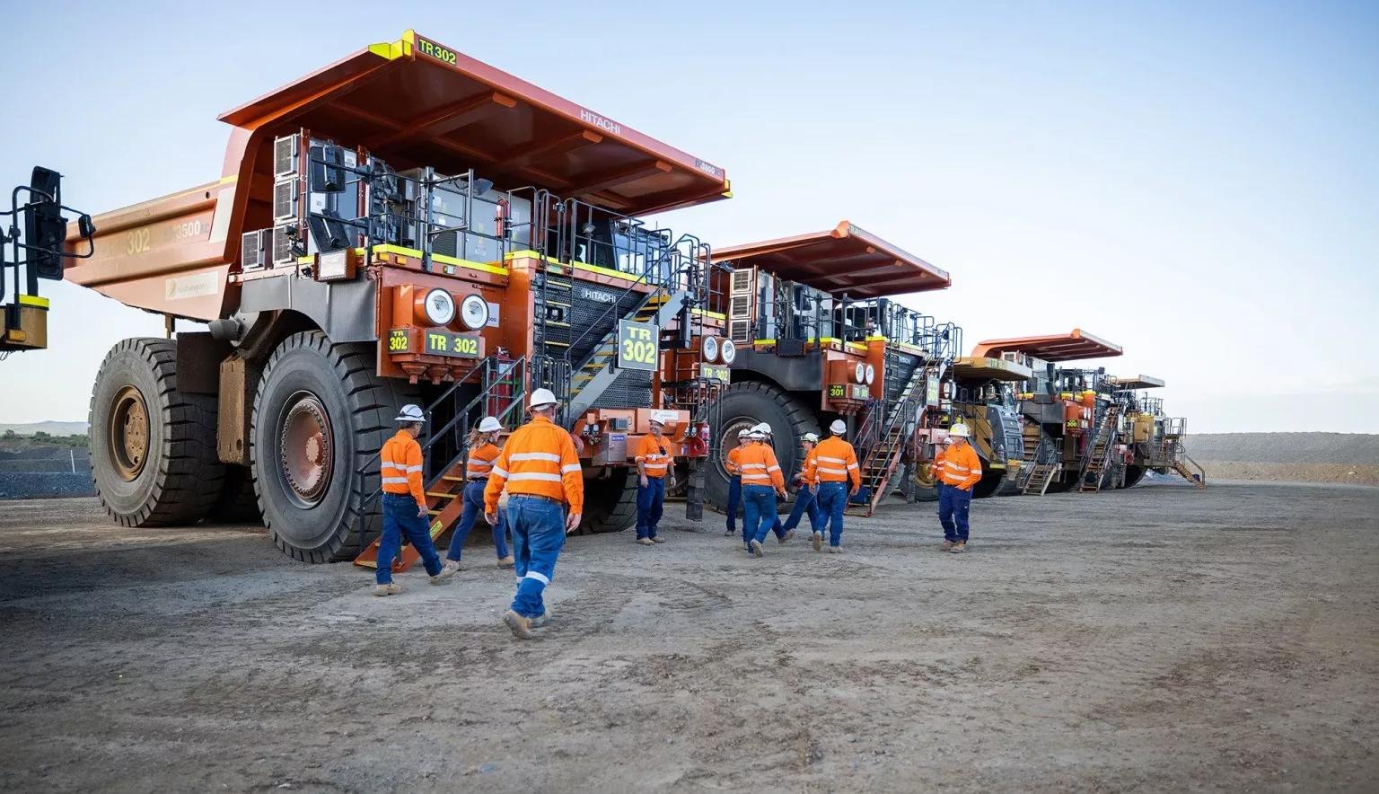 Mine site crew in hi-vis and hard hats walking past a line of parked haul trucks at an Australian open-cut operation